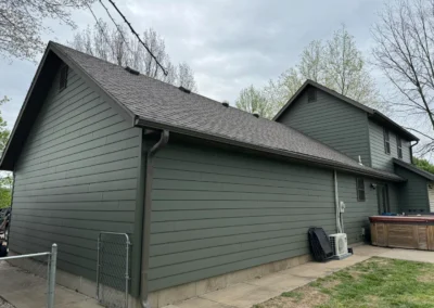 Side view of a modern green house with a gable roof, surrounded by trees, featuring an air conditioning unit and a hot tub.